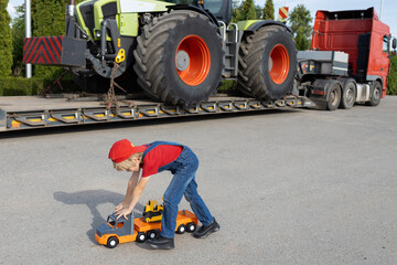 boy plays with toy cars in the parking lot next to a real big truck with a loaded tractor. The...