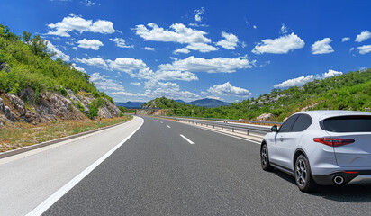 A white car drives along the highway against the backdrop of rocky mountains on a sunny day.