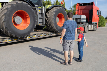 boy and a man, holding hands, examine a motorway car carrying a large tractor. The boy's interest in machines and their work. Father's Day