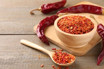 Aromatic spice. Red chili pepper flakes in bowl, spoon and pods on wooden table, closeup. Space for text