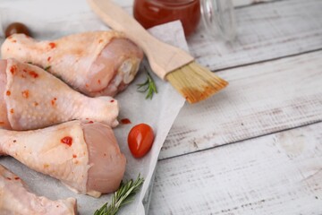 Raw marinated chicken drumsticks, rosemary and basting brush on white wooden table. Space for text
