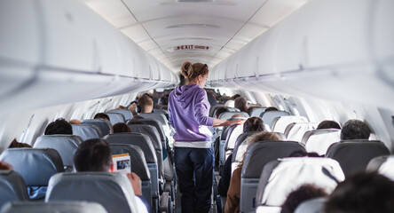 Interior of airplane with passengers on seats and female traveler walking the aisle. Commercial...