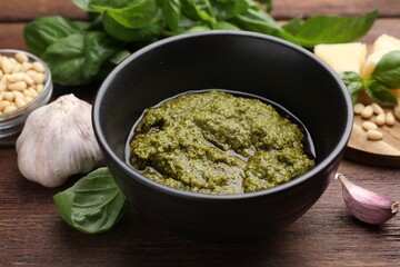Tasty pesto sauce in bowl, basil, garlic and pine nuts on wooden table, closeup