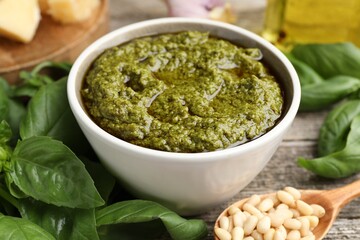 Tasty pesto sauce in bowl, pine nuts and basil on wooden table, closeup