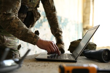 Military mission. Soldier in uniform using laptop at table inside abandoned building, closeup