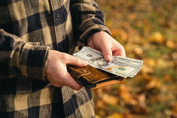 Leather wallet with dollar bills in male hands on a blurred nature background.