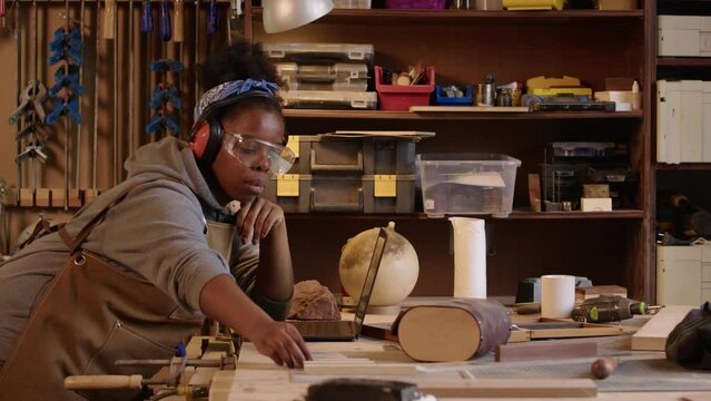 Medium shot of young African American female woodworker wearing safety glasses and earmuffs using laptop and examining wooden pieces while leaning on workbench at her workshop