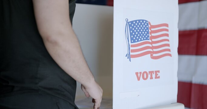 Close up from behind mixed race voter casting ballot at US election booth. Face not visible. US flag on wall in background