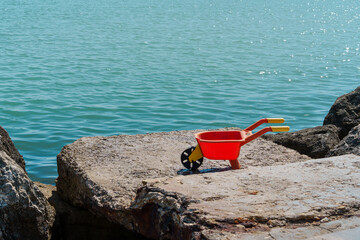 A red toy wheelbarrow on a rocky shore, against a sparkling sea backdrop, evokes childhood memories and the joy of seaside play