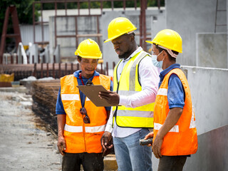 African male foreman and two Asian workers in safety vests and helmets stand by a building under...