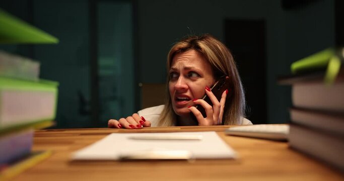 Scared woman speaks on phone asking for help. Terrified employee hides under table feeling afraid to stay alone in dark office in late evening during thunderstorm