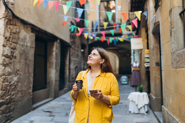 Happy cheerful young woman with paper coffee cup walking on city street checks her smartphone....