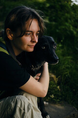 Young woman hugs with black mix breed dog in the middle of a green spring park. A dog with female owner rests outside on a summer sunny day in shadows of trees.