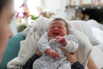 Newborn baby crying while being held, wearing a blue patterned onesie, with an expression of...