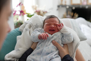 Newborn baby crying intensely while being held, wearing a blue patterned onesie, in a cozy home...
