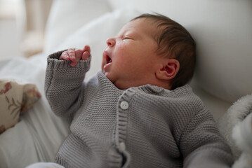 Newborn baby in a gray knitted cardigan and white pants, lying on a soft bed, with a slight smile...