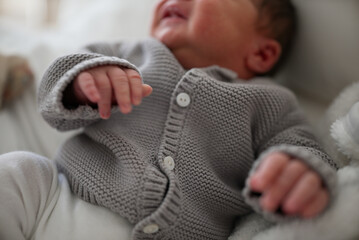Newborn baby in a gray knitted cardigan and white pants, lying on a soft bed, eyes closed, looking...