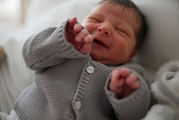 Newborn baby smiling while wearing a gray knitted sweater, lying on a white blanket. The baby has...