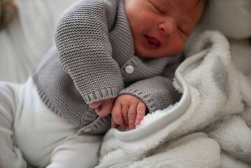Newborn baby with dark hair in a gray knitted sweater, lying on a white blanket, stretching and...