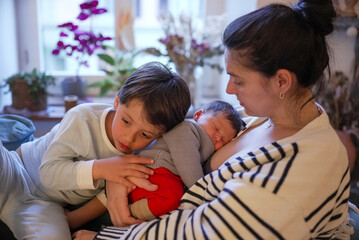 Mother holding her newborn while the older sibling gently touches the baby, creating a heartwarming...