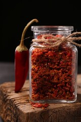 Chili pepper flakes in jar and pod on wooden board against black background, closeup