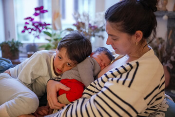 Older sibling gently interacting with a newborn baby while the mother lovingly watches. The room's...