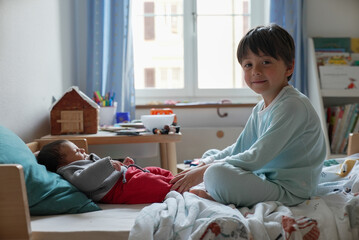Older brother in blue pajamas sits on a bed, smiling while watching over his newborn sibling who is...