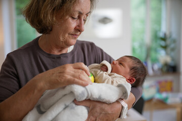 Grandmother gently holding her newborn grandchild wrapped in a white blanket, engaging the baby...