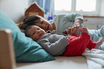 Older sibling lying down with newborn baby, gently holding the baby’s hand. The natural light...