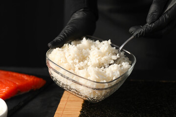 Chef in gloves taking cooked rice for sushi with spoon at dark table, closeup