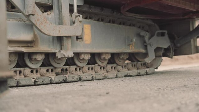 Riding caterpillar tracks asphalt paver video. Close-up view of the heavy-duty metal tracks in motion, leaving distinct imprints on the ground during road construction/paving operations.