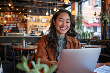 Fototapeta premium A smiling Latino woman using her laptop in a trendy cafe, showcasing the versatility and accessibility of remote work.