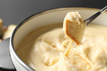 Dipping piece of bread into fondue pot with melted cheese on grey background, closeup