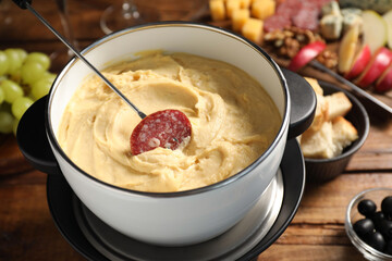 Fork with piece of sausage and melted cheese in fondue pot on wooden table, closeup