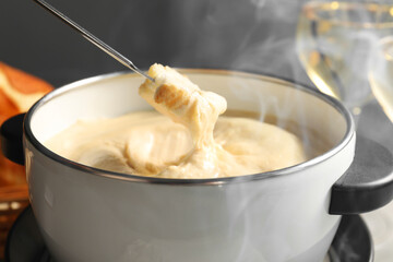 Dipping piece of bread into fondue pot with melted cheese at table, closeup