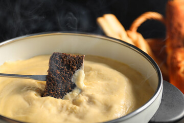 Dipping piece of bread into fondue pot with melted cheese on black background, closeup