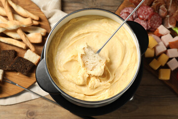 Dipping piece of bread into fondue pot with melted cheese at wooden table with snacks, flat lay