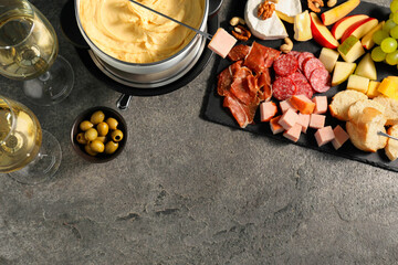 Fork with piece of ham, fondue pot with melted cheese, wine and snacks on grey textured table, flat...