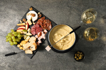 Fork with piece of ham, fondue pot with melted cheese, wine and snacks on grey textured table, flat...