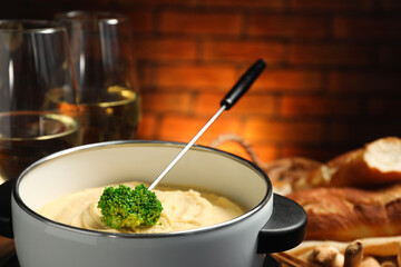 Fork with piece of broccoli, melted cheese in fondue pot, wine and snacks on blurred background,...