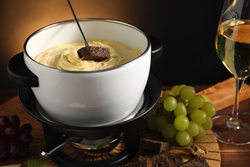Dipping piece of bread into fondue pot with melted cheese at wooden table with snacks, closeup