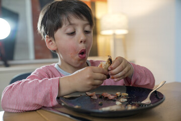 Close-up of a young boy in a pink striped shirt, concentrating on eating a slice of pizza. The...