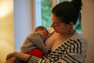 Mother in a striped sweater holds her sleeping newborn dressed in a grey sweater and red pants. The...