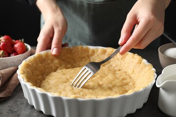 Shortcrust pastry. Woman making holes in raw dough with fork at grey table, closeup