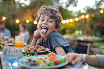 Lighthearted image of a young boy at a table outdoors, savoring grilled food at a family garden party, while sticking out his tongue in a playful manner.
