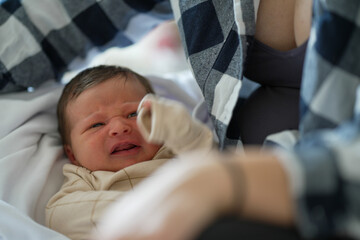 Close-up of a baby in beige clothes, lying down and looking slightly upset, with the mother's arms framing the scene. The baby&rsquo;s expression and the mother&rsquo;s presence convey a mix of emotions