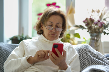 An elderly woman with glasses and a white sweater sits on a couch, intently using her smartphone....