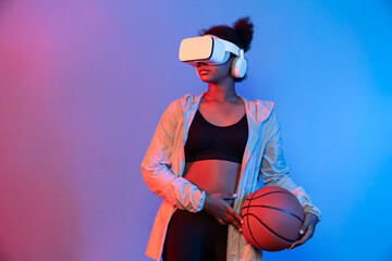 Young African woman playing game in basketball player wearing VR bouncing pose isolated blue neon...