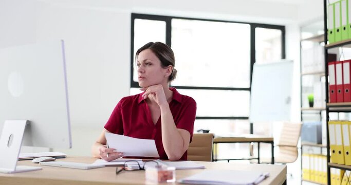 Woman worker looks at monitor screen with chin on hand hesitating at workplace in modern office. Female person thinks about correct task solution slow motion