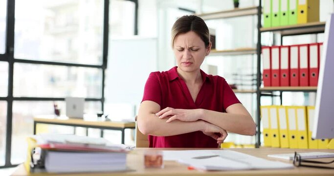 Woman office employee itches skin in area of arms sitting at table in light premise. Annoyed female feels skin discomfort due allergies irritation slow motion